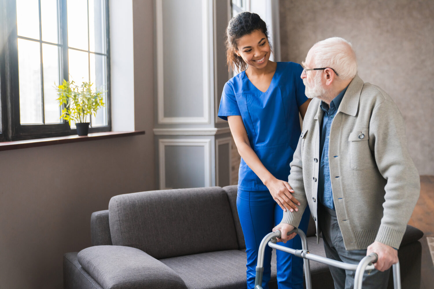 Portrait of an african young nurse helping old elderly disable man grandfather to walk using walker equipment in the bedroom. Senior patient of nursing home moving with walking frame and nurse support