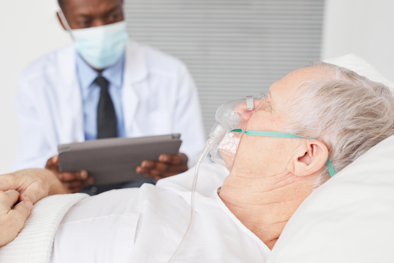 Side view close up of senior man lying in hospital bed with oxygen mask and African-American doctor in background, copy space